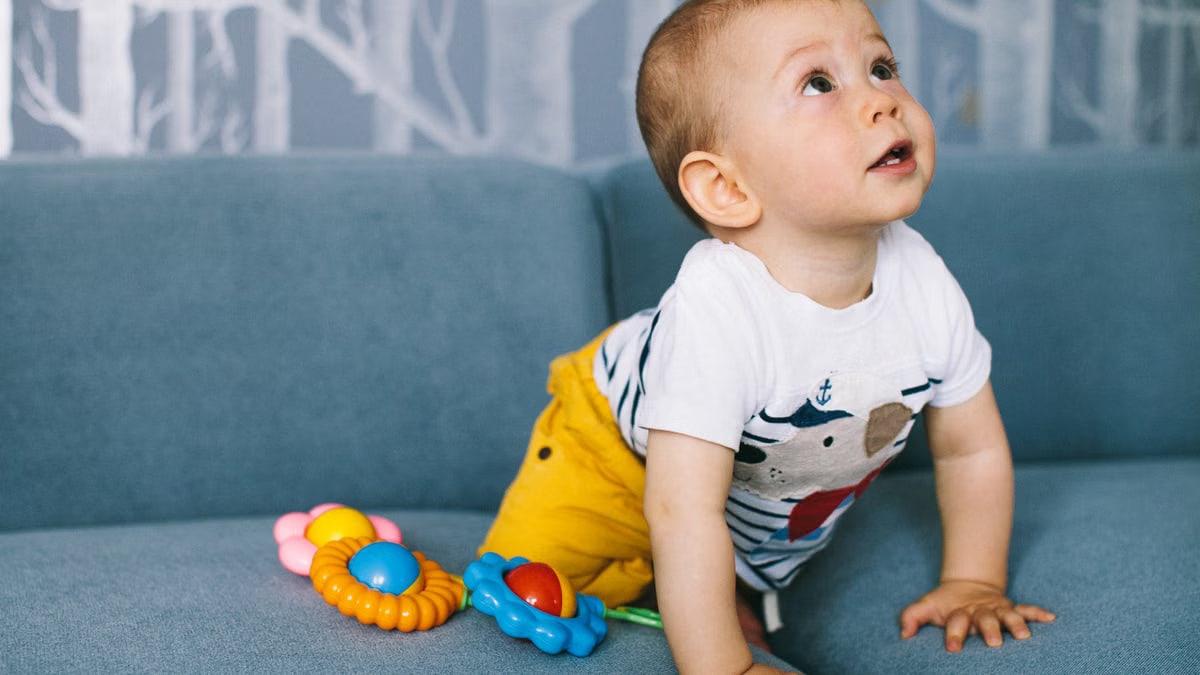 Adorable baby boy crawling on a blue sofa, surrounded by colorful toys, showcasing curiosity and innocence.